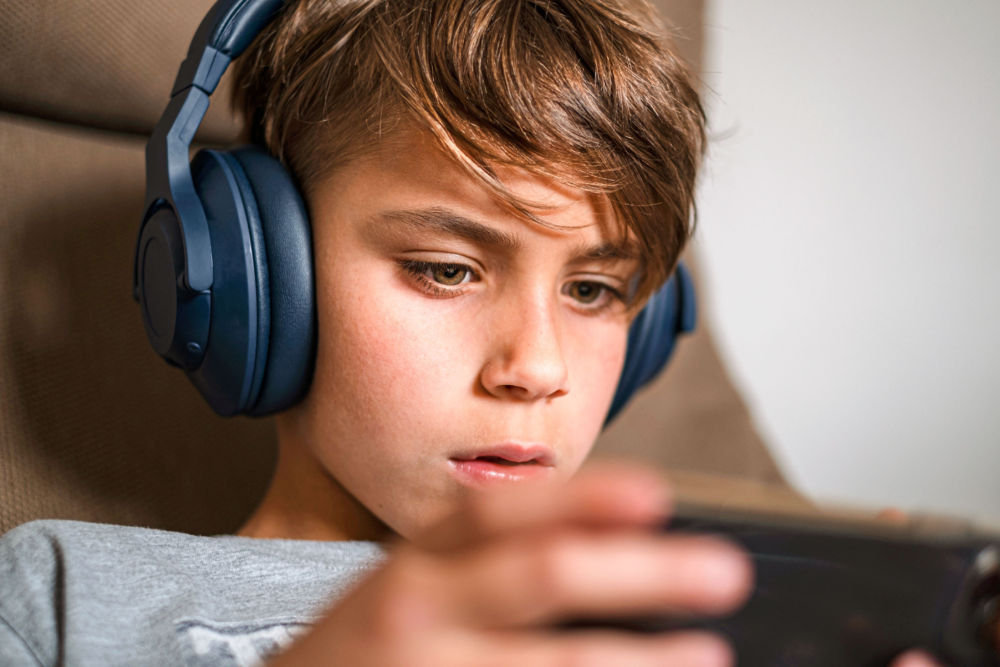 Nahaufnahme eines Jungen mit großen, dunkelblauen Kopfhörern, der hochkonzentriert auf ein mobiles Gerät in seinen Händen blickt. Sein Gesichtsausdruck ist ernst und völlig in die digitale Aktivität versunken. Das Bild illustriert Gaming-Kultur bei Kindern, die Nutzung von Audio-Inhalten zur Abschirmung und die starke Anziehungskraft von Videospielen. Es symbolisiert Themen wie Reizüberflutung, die Notwendigkeit von Medienpausen und die Beratung des Psychosozialen Gesundheitszentrums zur Prävention von Gaming-Sucht und zur Förderung einer gesunden digitalen Balance.