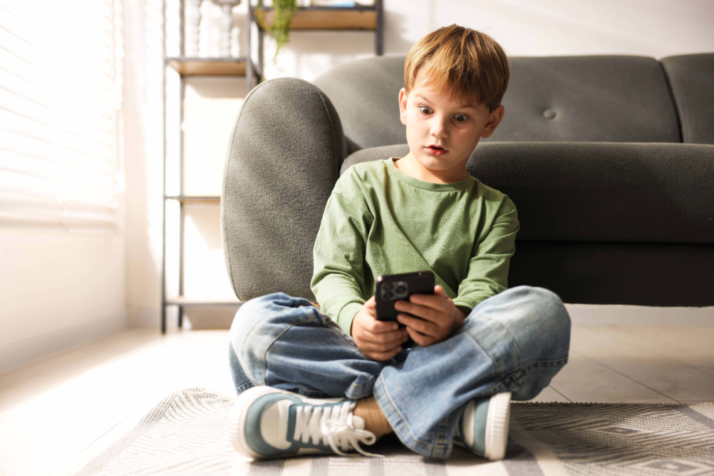 Ein Junge sitzt allein im Schneidersitz auf einem Teppich vor einem Sofa und blickt konzentriert auf ein Smartphone in seinen Händen. Das Bild illustriert die frühe Konfrontation mit digitalen Inhalten, die Alleinnutzung von Medien im Kinderzimmer und die Faszination für mobile Endgeräte. Es symbolisiert die Notwendigkeit von Kinderschutzeinstellungen, die Beratung zur Medienkompetenz durch das Psychosoziale Gesundheitszentrum und die Begleitung von Kindern in einer vernetzten Welt.