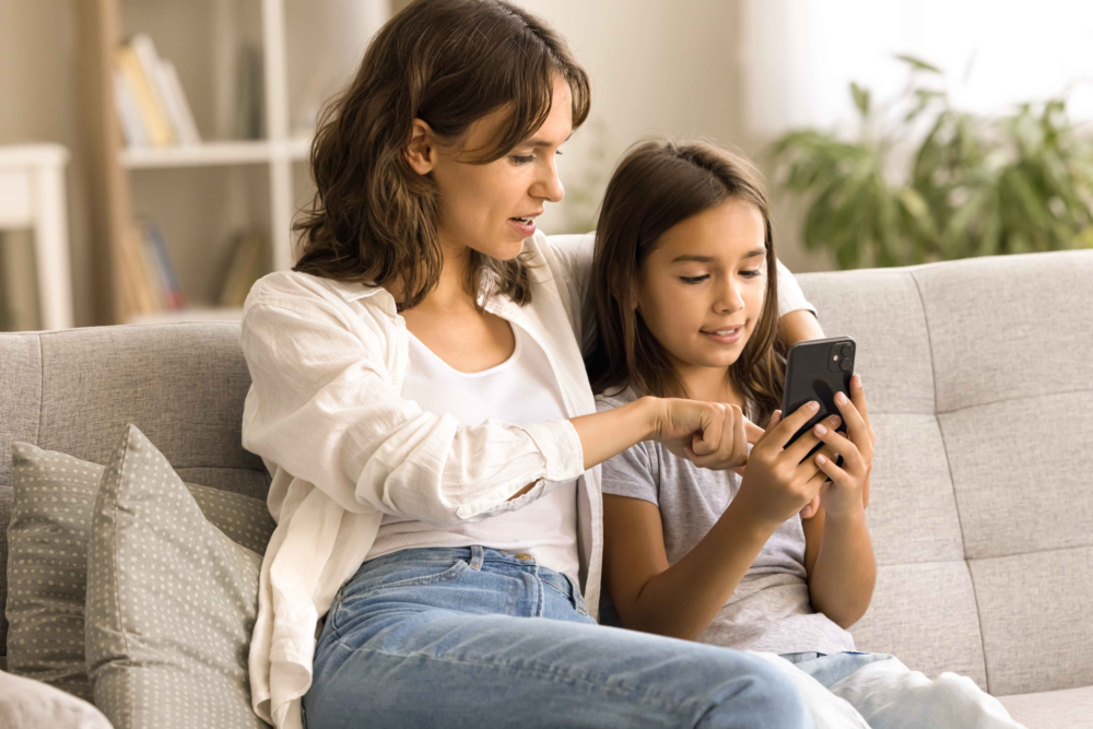 Eine Mutter und ihre Tochter sitzen eng beieinander auf einem hellen Sofa. Die Mutter zeigt der Tochter etwas auf einem Smartphone und erklärt es ihr mit zugewandter Mimik, während das Mädchen aufmerksam auf das Display blickt. Das Bild illustriert aktive Medienbegleitung, das gemeinsame Erkunden digitaler Inhalte und eine vertrauensvolle Mutter-Kind-Interaktion. Es symbolisiert die Förderung von Medienkompetenz in der Familie und die präventive Beratung des Psychosozialen Gesundheitszentrums zum sicheren Umgang mit dem Internet.