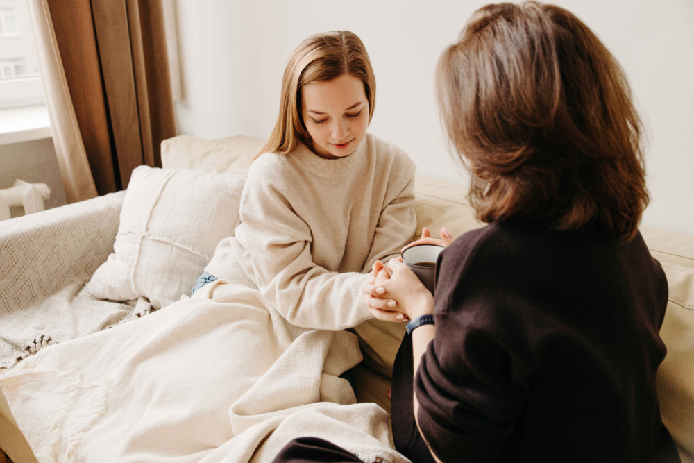 Nahaufnahme einer einfühlsamen Beratungssituation: Eine Frau hält tröstend die Hände einer jungen Frau, die erschöpft, aber sicher auf einem Sofa sitzt. Beide sind in eine Decke gehüllt, was eine Atmosphäre von maximaler Geborgenheit und Schutz vermittelt. Das Bild illustriert Krisenintervention, emotionale Unterstützung bei Erschöpfung oder Trauer und die tiefe menschliche Ebene der psychosozialen Arbeit. Es symbolisiert den sicheren Hafen, den das Psychosoziale Gesundheitszentrum für Menschen in seelischer Not bietet.“