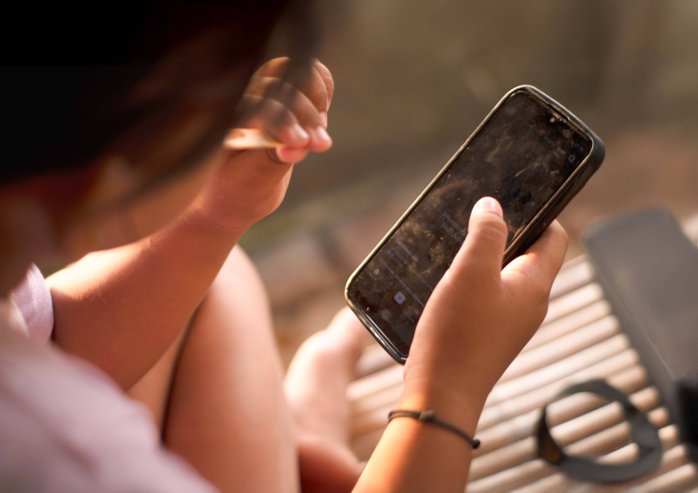 Nahaufnahme aus der Vogelperspektive auf ein Kind, das ein Smartphone in beiden Händen hält und konzentriert bedient. Der Fokus liegt auf den kleinen Händen und dem leuchtenden Display, während der Hintergrund in warmes Sonnenlicht getaucht ist. Das Bild illustriert die frühe Mediennutzung, die intuitive Bedienung von Mobilgeräten durch Kinder und die Herausforderung der digitalen Balance im Alltag. Es symbolisiert Themen wie App-Nutzung, Online-Sicherheit für Minderjährige und die pädagogische Begleitung der digitalen Kindheit.
