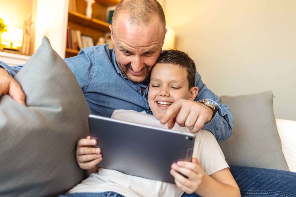 Ein Vater und sein Sohn sitzen eng zusammen auf einem Sofa und bedienen gemeinsam ein Tablet. Beide lachen herzlich, während der Vater mit dem Finger auf den Bildschirm zeigt. Das Bild illustriert positive Medienerziehung, den Spaß an gemeinsamer digitaler Freizeitgestaltung und eine starke Vater-Kind-Bindung. Es symbolisiert die Begleitung von Kindern im Internet, das Teilen von Interessen und die Förderung von Medienkompetenz in einem geschützten, familiären Rahmen.