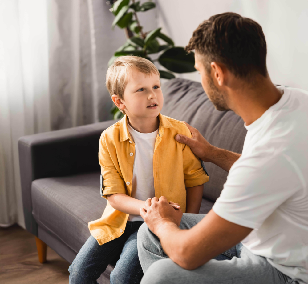 Ein Vater sitzt seinem sichtlich traurigen Sohn auf einem grauen Sofa gegenüber. Er hält die Hände des Jungen fest in seinen und hat ihm tröstend eine Hand auf die Schulter gelegt, während sie ein ernstes, aber liebevolles Gespräch führen. Das Bild illustriert väterliche Empathie, die Krisenbewältigung in der Familie und den Aufbau von Vertrauen in schwierigen Lebensphasen. Es symbolisiert Sicherheit, männliche Bezugspersonen als emotionale Stütze und die professionelle Begleitung durch das Psychosoziale Gesundheitszentrum (PSGZ) bei Verhaltensauffälligkeiten oder Sorgen.