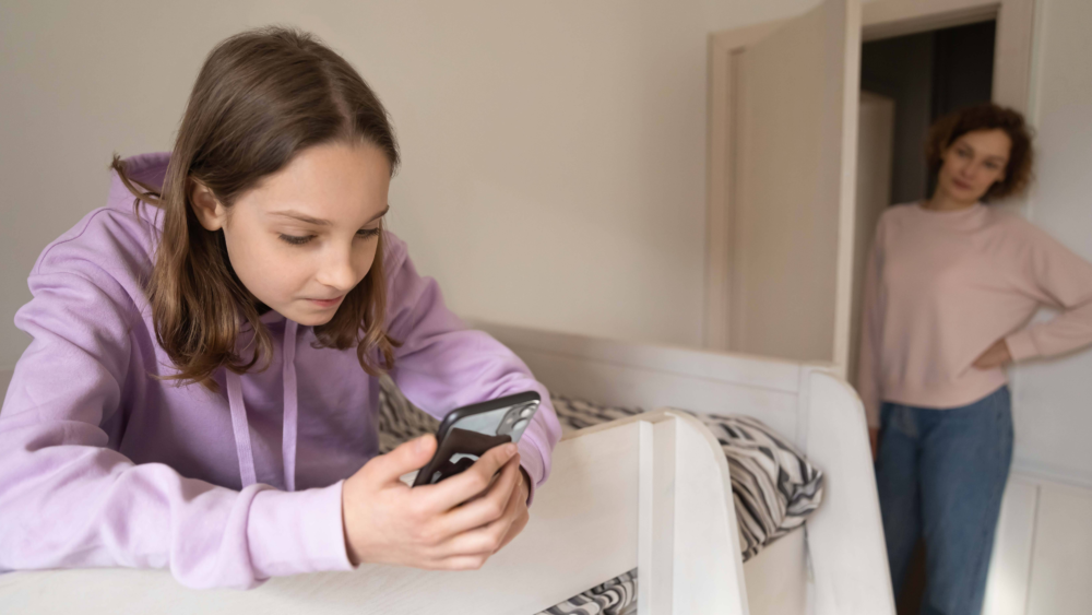 Ein junges Mädchen in einem lila Hoodie liegt auf ihrem Bett und starrt gebannt auf ihr Smartphone. Im unscharfen Hintergrund steht eine Frau im Türrahmen und beobachtet die Szene mit einem besorgten und skeptischen Gesichtsausdruck. Das Bild illustriert Konflikte um die Handynutzung bei Teenagern, die emotionale Entfremdung durch übermäßigen Medienkonsum und die elterliche Sorge um die digitale Sicherheit. Es symbolisiert die Kluft zwischen der digitalen Welt der Jugendlichen und der analogen Realität der Eltern.