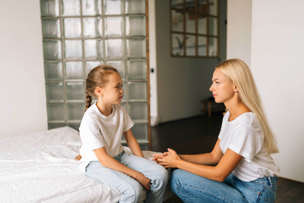 Eine Frau in einer weißen Bluse kniet einfühlsam vor einem kleinen Mädchen, das traurig auf einem Bett sitzt. Die Frau hält sanft die Hände des Kindes und sucht den Blickkontakt auf Augenhöhe. Das Bild illustriert eine professionelle psychosoziale Beratung, die Begleitung in schwierigen Lebensphasen und das Thema Kinderschutz. Es symbolisiert Empathie, das Schaffen eines sicheren Raums für Gespräche und die emotionale Unterstützung bei Sorgen oder Traumata.