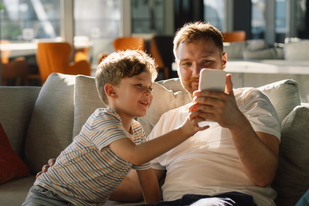 Ein Mann und ein kleiner Junge sitzen zusammen auf einem grauen Sofa in einem lichtdurchfluteten Wohnzimmer. Der Mann hält ein weißes Smartphone hoch, auf das beide lächelnd und interessiert blicken. Die Szene ist von warmem Sonnenlicht geprägt, das eine freundliche Atmosphäre schafft. Das Bild illustriert das gemeinsame Anschauen von Inhalten, die Begleitung des Kindes bei der Mediennutzung durch den Vater und einen Moment der Nähe und Freude. Es symbolisiert positive Vorbildfunktion, digitalen Austausch in der Familie und die Stärkung der Bindung durch geteilte Erlebnisse.