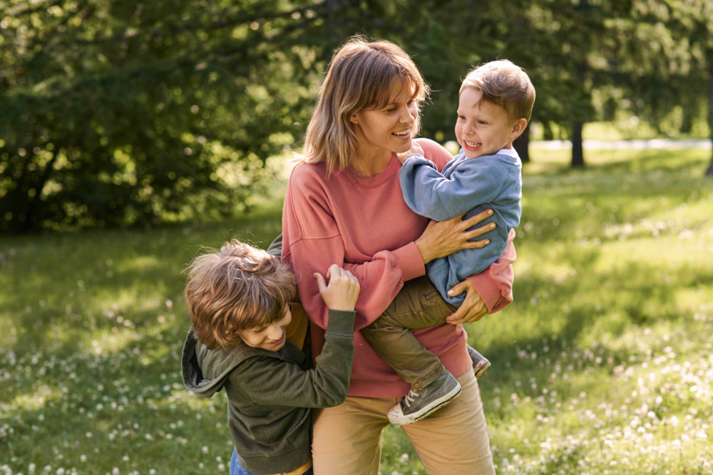 Eine lachende Mutter steht auf einer sonnigen, grünen Wiese und hält ein kleines Kind spielerisch im Arm, während ein zweites Kind fröhlich an ihrer Seite springt. Alle drei wirken glücklich und genießen die gemeinsame Zeit unter Bäumen. Das Bild illustriert aktive Freizeitgestaltung in der Natur, die emotionale Bindung zwischen Mutter und Kindern und die Bedeutung von Bewegung an der frischen Luft. Es symbolisiert Lebensfreude, familiären Zusammenhalt und einen gesunden, naturnahen Lebensstil abseits digitaler Ablenkungen.