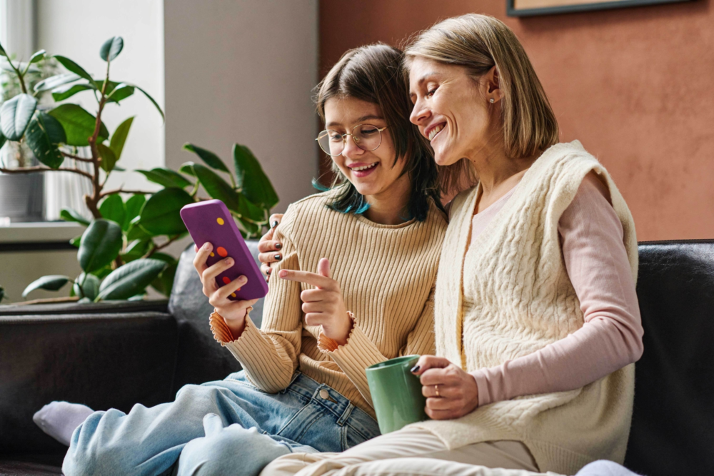 Eine Frau und ein Mädchen sitzen entspannt nebeneinander auf einem dunklen Sofa. Das Mädchen hält ein Smartphone in einer lila Hülle und zeigt der lächelnden Frau aufmerksam einen Inhalt auf dem Bildschirm. Beide wirken fröhlich und befinden sich in einem aktiven Austausch. Im Hintergrund ist eine große grüne Zimmerpflanze zu sehen. Das Bild illustriert gemeinsame Mediennutzung, das Mentoring durch Erwachsene und eine vertrauensvolle Beziehung. Es symbolisiert digitale Teilhabe, den Abbau von Berührungsängsten gegenüber Technik und die Stärkung der Bindung durch geteilte digitale Erlebnisse im häuslichen Umfeld.