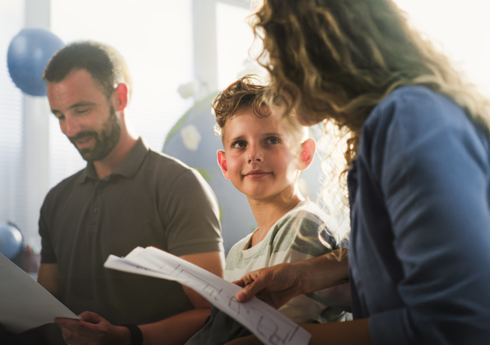 Eine Beraterin mit lockigem Haar sitzt mit einem Paar und deren Sohn an einem hellen Tisch. Sie hält Dokumente in der Hand und erklärt diese ruhig, während der Junge aufmerksam zuhört und die Eltern die Unterlagen prüfen. Das Bild illustriert eine professionelle Familienberatung, das gemeinsame Erarbeiten von Lösungen und die transparente Informationsvermittlung. Es symbolisiert Mitspracherecht für Kinder, professionelle Unterstützung bei Erziehungsfragen und die Arbeit des Psychosozialen Gesundheitszentrums zur Stärkung der familiären Kooperation und Kommunikation.