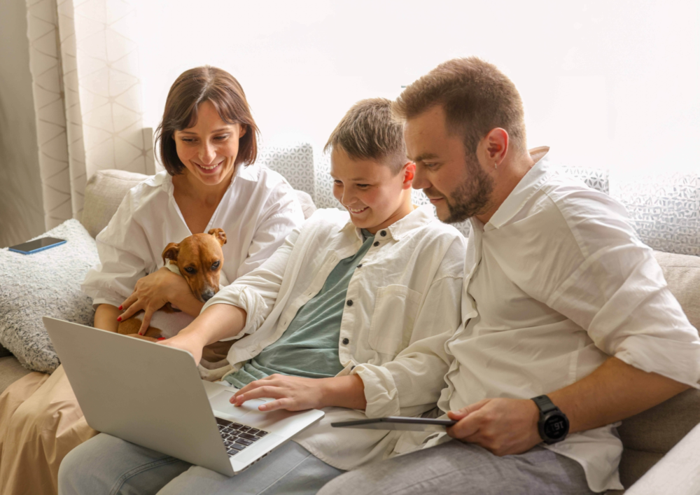 Eine dreiköpfige Familie sitzt entspannt auf einem hellen Sofa und nutzt gemeinsam einen Laptop. Der Sohn bedient die Tastatur, während Vater und Mutter (mit einem kleinen Hund auf dem Schoß) interessiert und lächelnd auf den Bildschirm schauen. Das Bild illustriert familienzentrierte Mediennutzung, das gemeinsame Lernen am Computer und eine positive Begleitung durch die Eltern. Es symbolisiert Zusammenhalt, digitale Teilhabe für alle Generationen und die Beratung des Psychosozialen Gesundheitszentrums zur Förderung einer gesunden und verbindenden Familienkommunikation im digitalen Zeitalter.